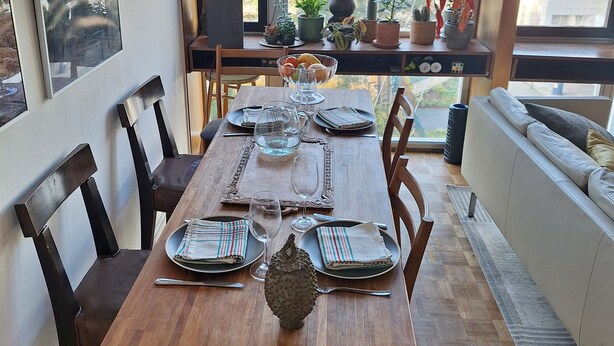 A dining area with a wooden table set for four, surrounded by chairs. There are potted plants on a window sill, and a view of buildings outside.