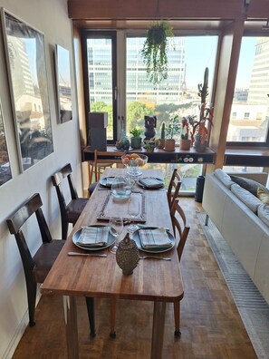 Un coin repas avec une table en bois dressée pour quatre personnes, entourée de chaises. Des plantes en pot sont posées sur le rebord d’une fenêtre, et on peut admirer la vue sur les bâtiments à l’extérieur.