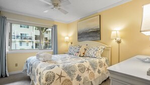 A bedroom with a bed, a ceiling fan, a window with curtains, a nightstand, and a wall-mounted lamp. - Tanglewood 101 - South Forest Beach 1 Block From the Ocean (Hilton Head Island)