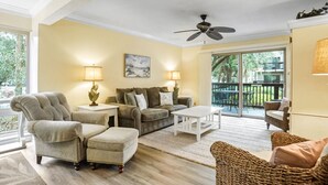 A spacious living room with a sofa, armchairs, and a coffee table. There is a ceiling fan, a painting on the wall, and a view of the outdoors through a sliding glass door. - Tanglewood 101 - South Forest Beach 1 Block From the Ocean (Hilton Head Island)