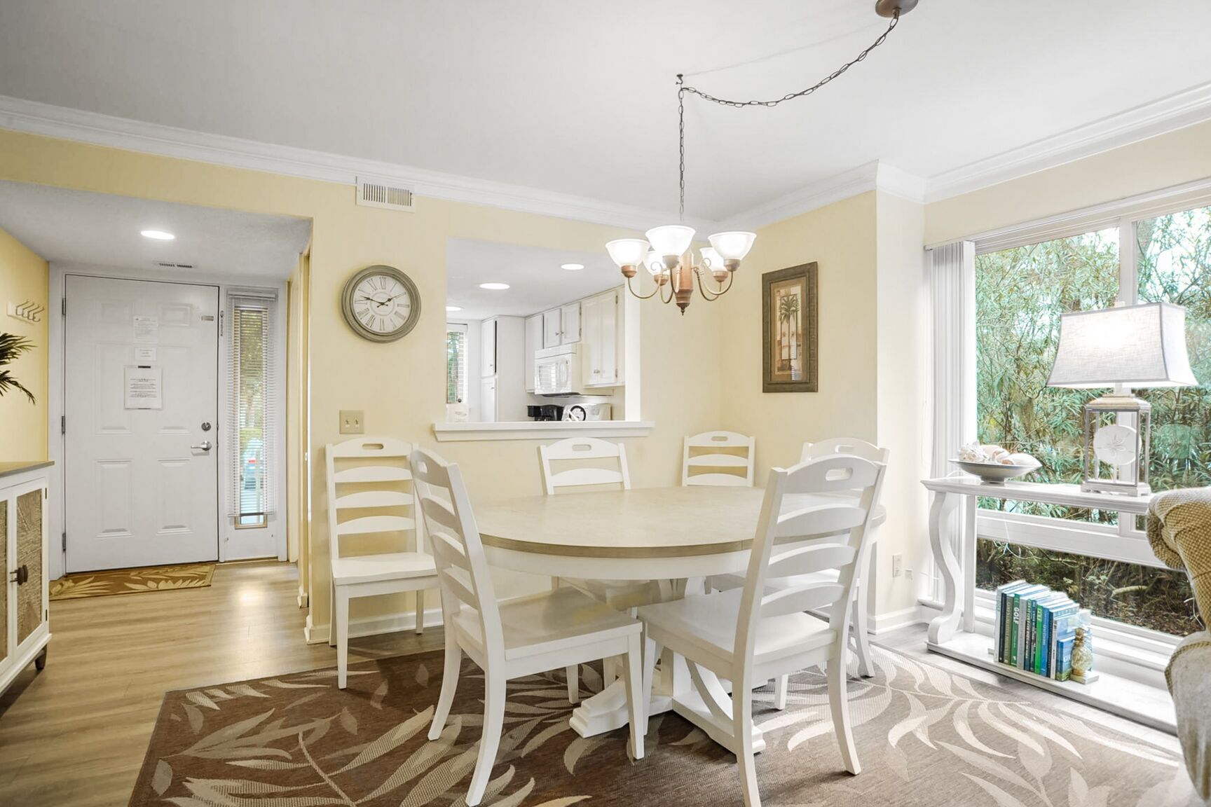 A dining area with a round table and white chairs, a kitchen in the background, and a window with a view of greenery.