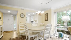 A dining area with a round table and white chairs, a kitchen in the background, and a window with a view of greenery. - Tanglewood 101 - South Forest Beach 1 Block From the Ocean (Hilton Head Island)