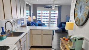 A compact kitchen with a central island, a dining area with a blue sofa, and a large wall clock. - Oceanview Midtown Condo with Pool! SS66500 (Ocean City)