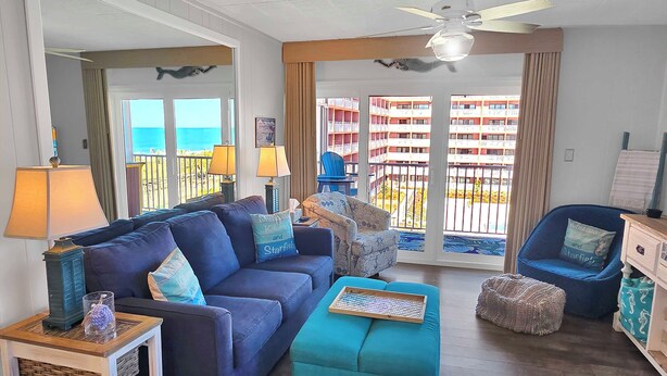 A living room with a sofa, ottoman, and chairs, a ceiling fan, and a view of the ocean through large windows. - Oceanview Midtown Condo with Pool! SS66500 (Ocean City)