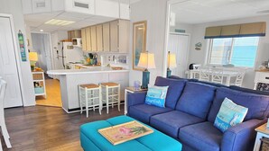 A modern living room with a blue sofa, a matching ottoman, and a kitchen in the background. - Oceanview Midtown Condo with Pool! SS66500 (Ocean City)