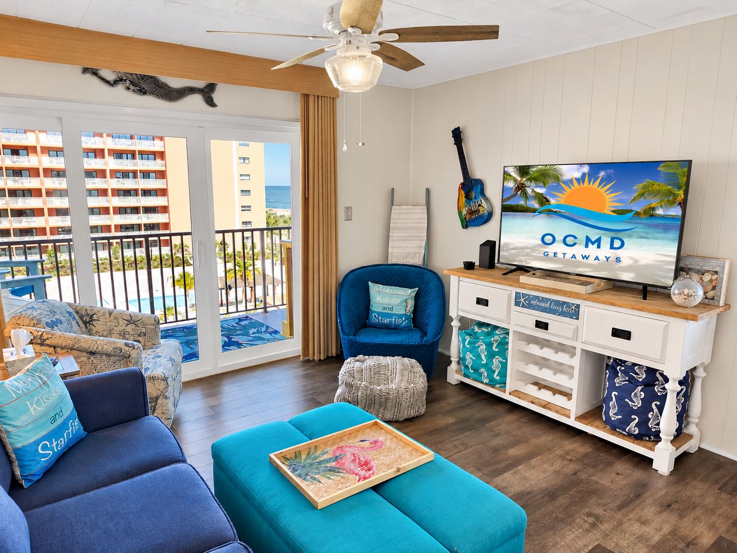 A living room with a television displaying 'OCMD GETAWAYS,' a blue armchair, a sofa, a wooden coffee table, and a view of the ocean through a sliding glass door.
