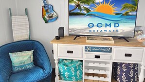 A room with a TV displaying 'OCMD GETAWAYS' on a beach scene, a guitar, a blue chair, and a white cabinet with drawers. - Oceanview Midtown Condo with Pool! SS66500 (Ocean City)