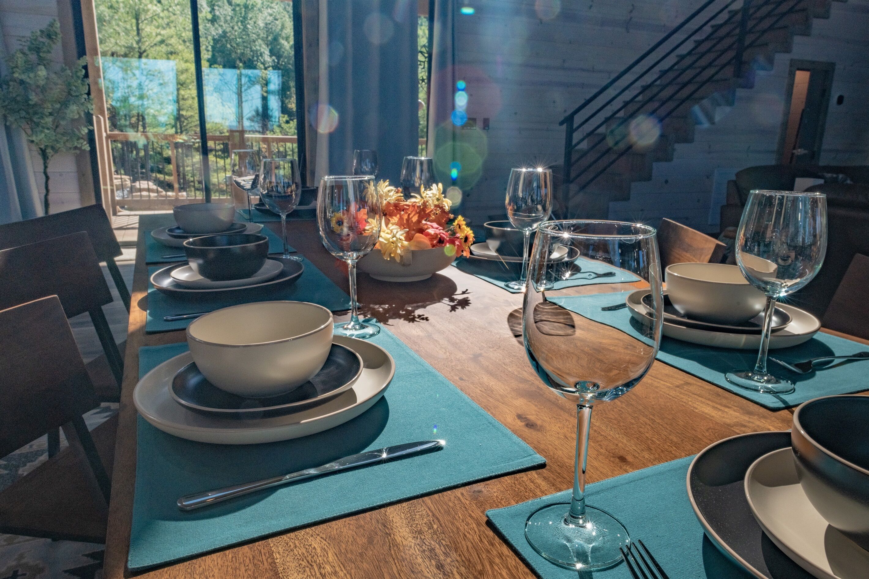 Uma mesa de jantar posta com tigelas, pratos e copos de vinho, com vista para árvores e uma escadaria ao fundo.
