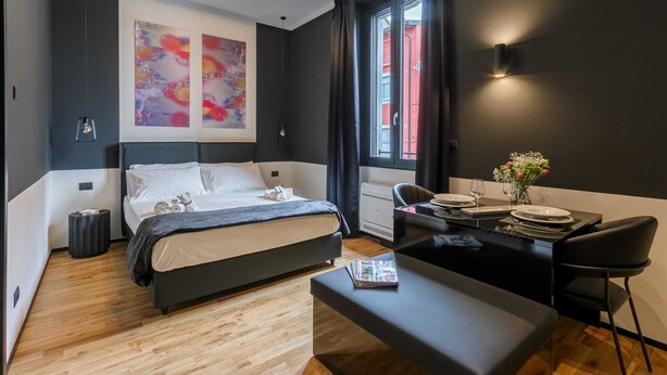 Ett modernt hotellrum med en stor säng, ett skrivbord med en stol, ett litet bord med en vas med blommor och konstverk på väggen.