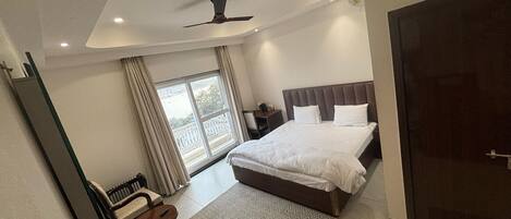 Una habitación de hotel moderna con una cama grande, un escritorio y una silla. Hay una ventana con cortinas, un ventilador de techo y una puerta.