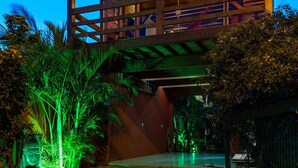 Un edificio de madera de dos pisos con balcón, rodeado de vegetación tropical y un camino pavimentado.