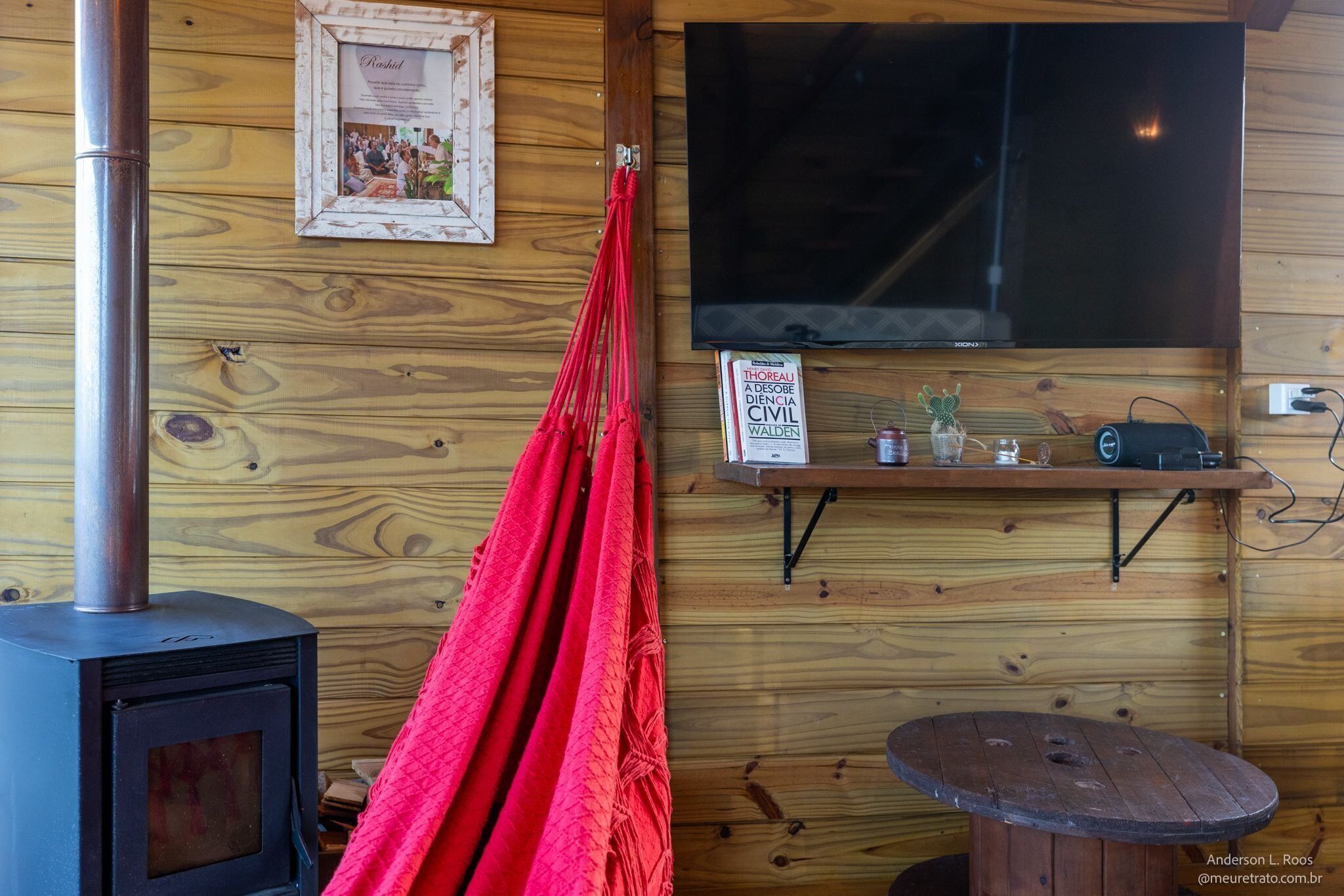 Een houten blokhut van binnen met een houtkachel, een hangmat, een wandtelevisie en een klein plankje met spullen.