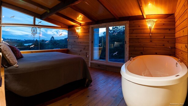Un dormitorio de cabaña de madera con una cama grande, una bañera y vistas del paisaje a través de las ventanas.