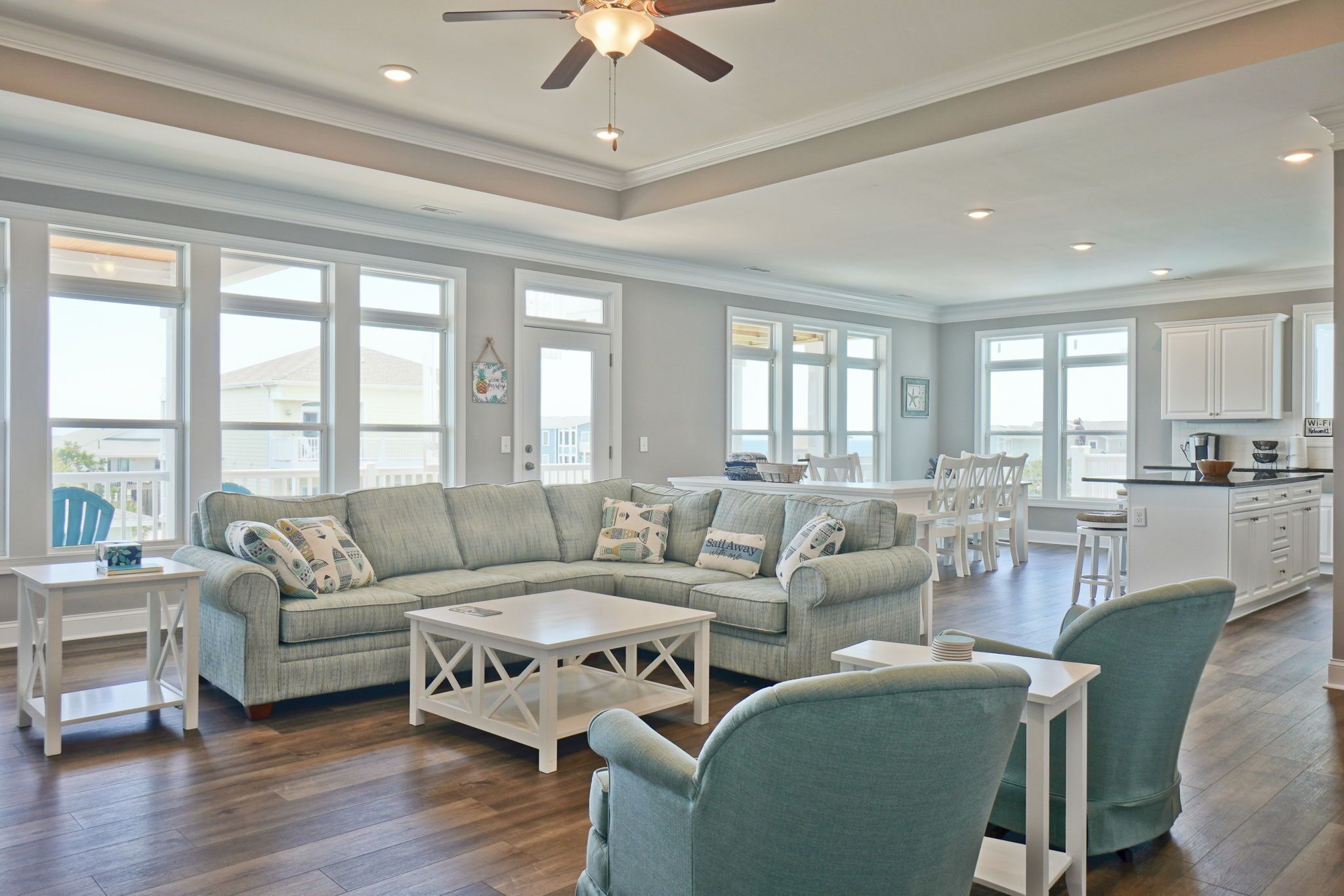A spacious living room with a sectional sofa, a dining area with white chairs, a kitchen with white cabinets, and large windows providing natural light.
