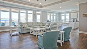 Een ruime woonkamer met een modulaire bank, een bijzettafeltje en teal-kleurige stoelen. Er is een keuken met witte kasten en een plafondventilator.
