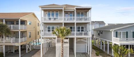 A multi-story beachfront house with a balcony, a palm tree, and a clear sky.