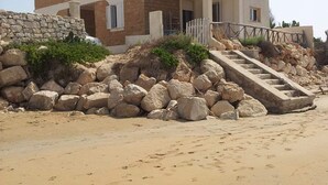 A modern beach house with a stone facade, a covered porch, and a stone staircase leading to the entrance. - Villa Sabbia Granelli (Pachino)