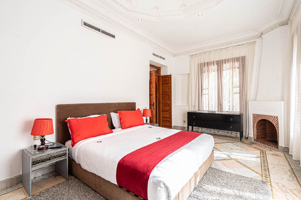 A bedroom with a large bed, red and white bedding, a nightstand with a lamp, a dresser, and a fireplace.