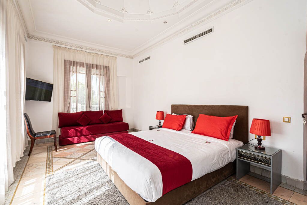 A bedroom with a large bed, red bedding, a red sofa, a TV, and a nightstand with a lamp.