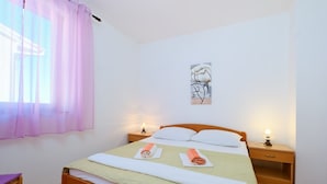 Ein ordentlich bezogenes Bett mit einer grünen Tagesdecke und zwei aufgerollten Handtüchern, ein Nachttisch mit einer Lampe sowie ein Fenster mit rosa Vorhängen.