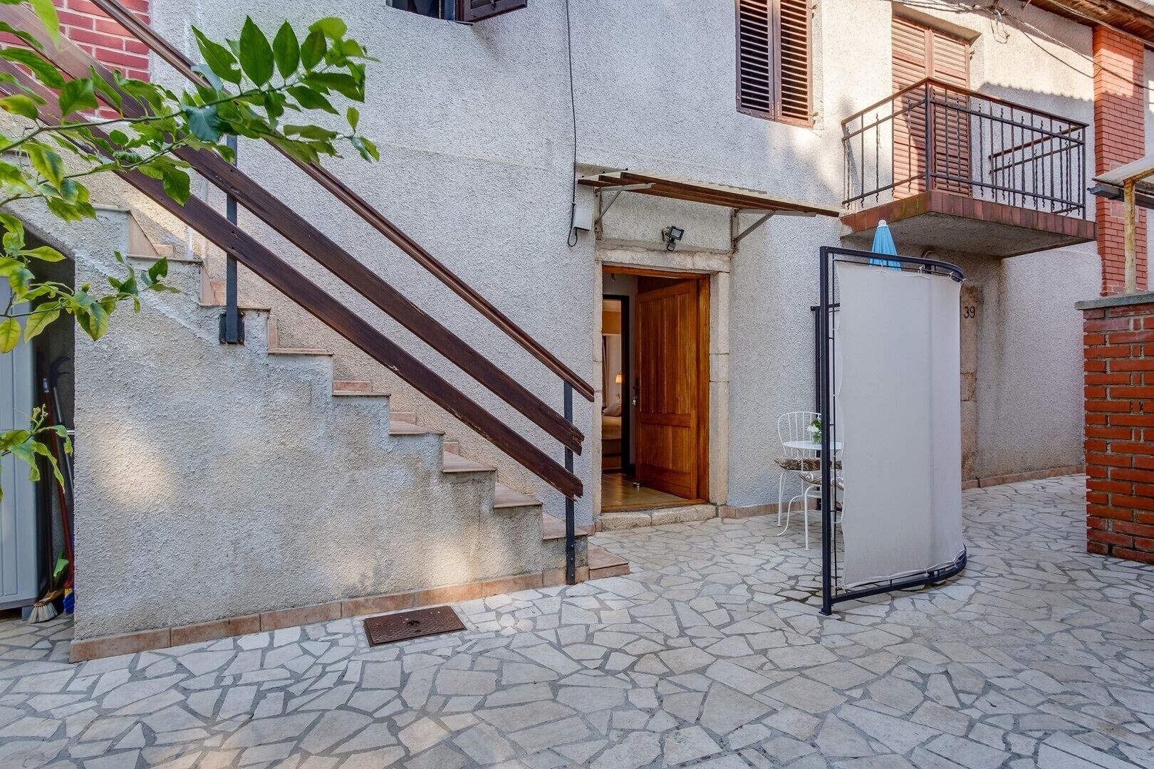 A courtyard with stone flooring, a wooden staircase, a white door, and a small table with a chair.