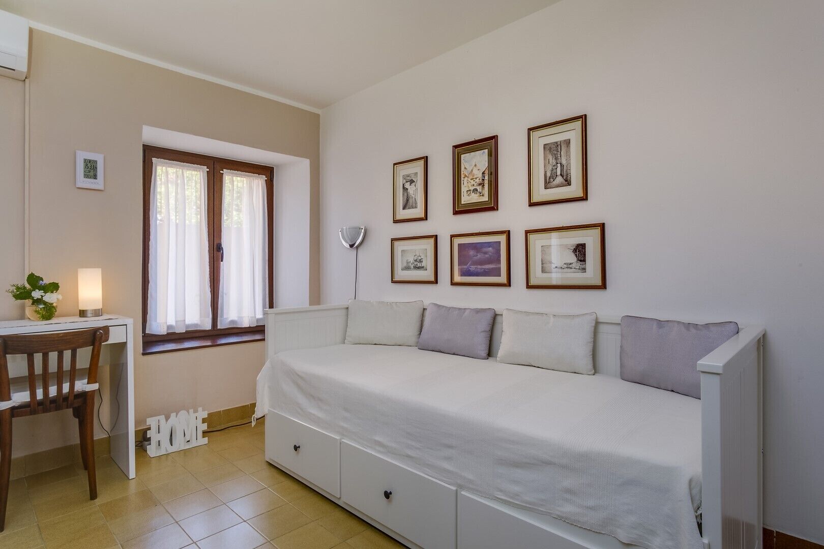 Una habitación pequeña y luminosa con una cama, un escritorio con una silla y una ventana con cortinas. Varias fotografías enmarcadas adornan la pared.