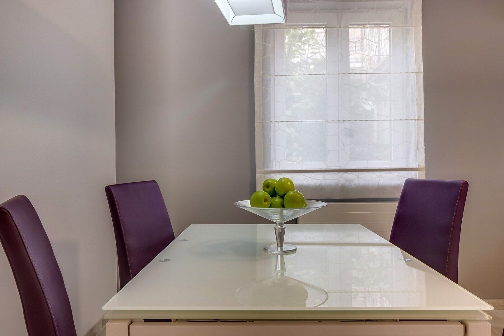 A dining table with a glass bowl of green apples, two purple chairs, and a window with blinds.