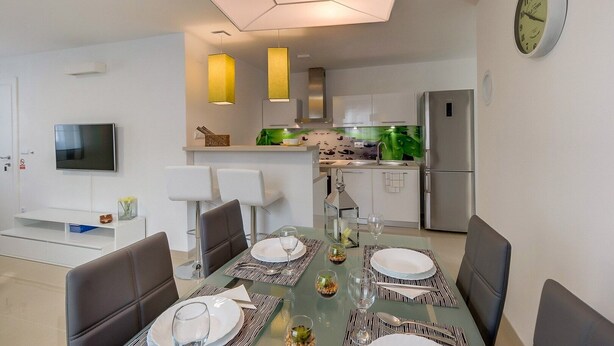 A modern kitchen with a built-in oven and a refrigerator. There is a dining area with a table set for four, and a TV mounted on the wall.