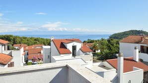 A coastal residential area with terracotta-tiled roofs, white buildings, and a clear view of the sea and hills in the background.