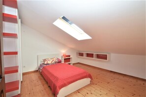 Une chambre avec un plafond en pente, un puits de lumière, un lit recouvert d’une couverture rouge, une table de chevet avec une lampe et des étagères intégrées.
