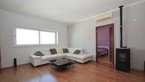 A modern living room with a white sofa, a glass coffee table, a black wood-burning stove, and a large window.