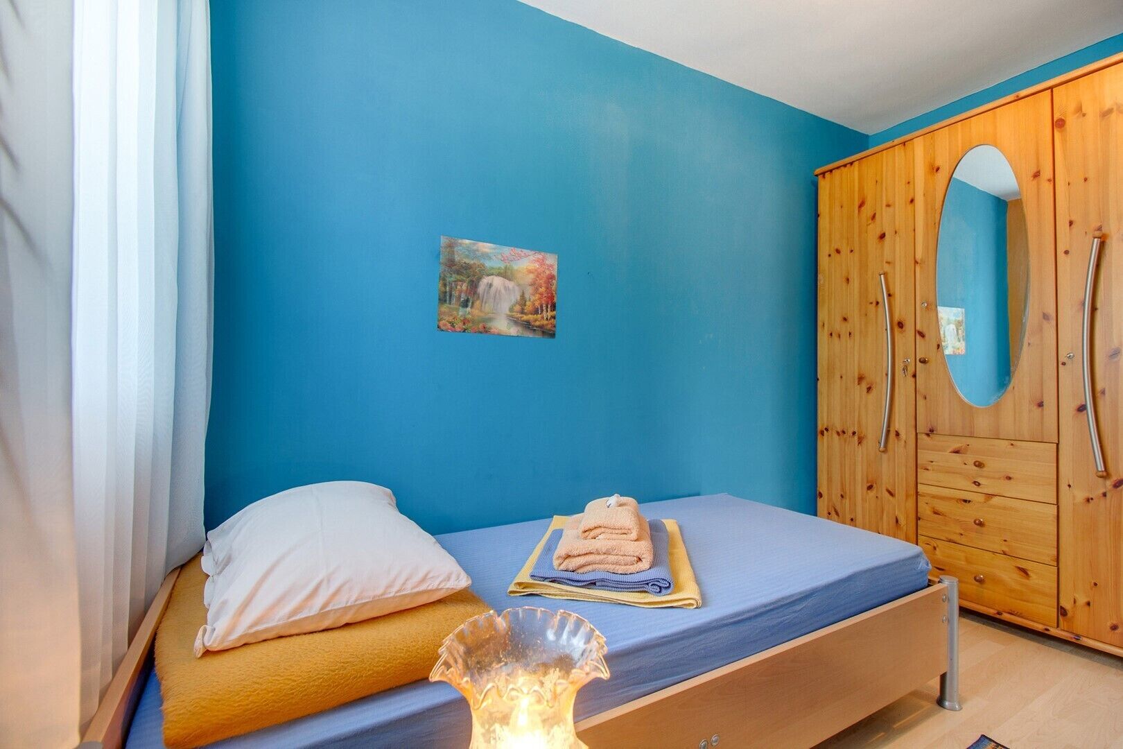 A bedroom with a blue wall, a wooden wardrobe with a mirror, a bed with a white pillow, and a small painting.