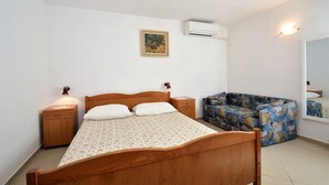 A bedroom with a wooden bed, a nightstand, a sofa, a painting, and an air conditioning unit. - Studio Rahela 3 in Mali Lošinj, Zagazinjine. Suitable for 2 people. (Mali Losinj)