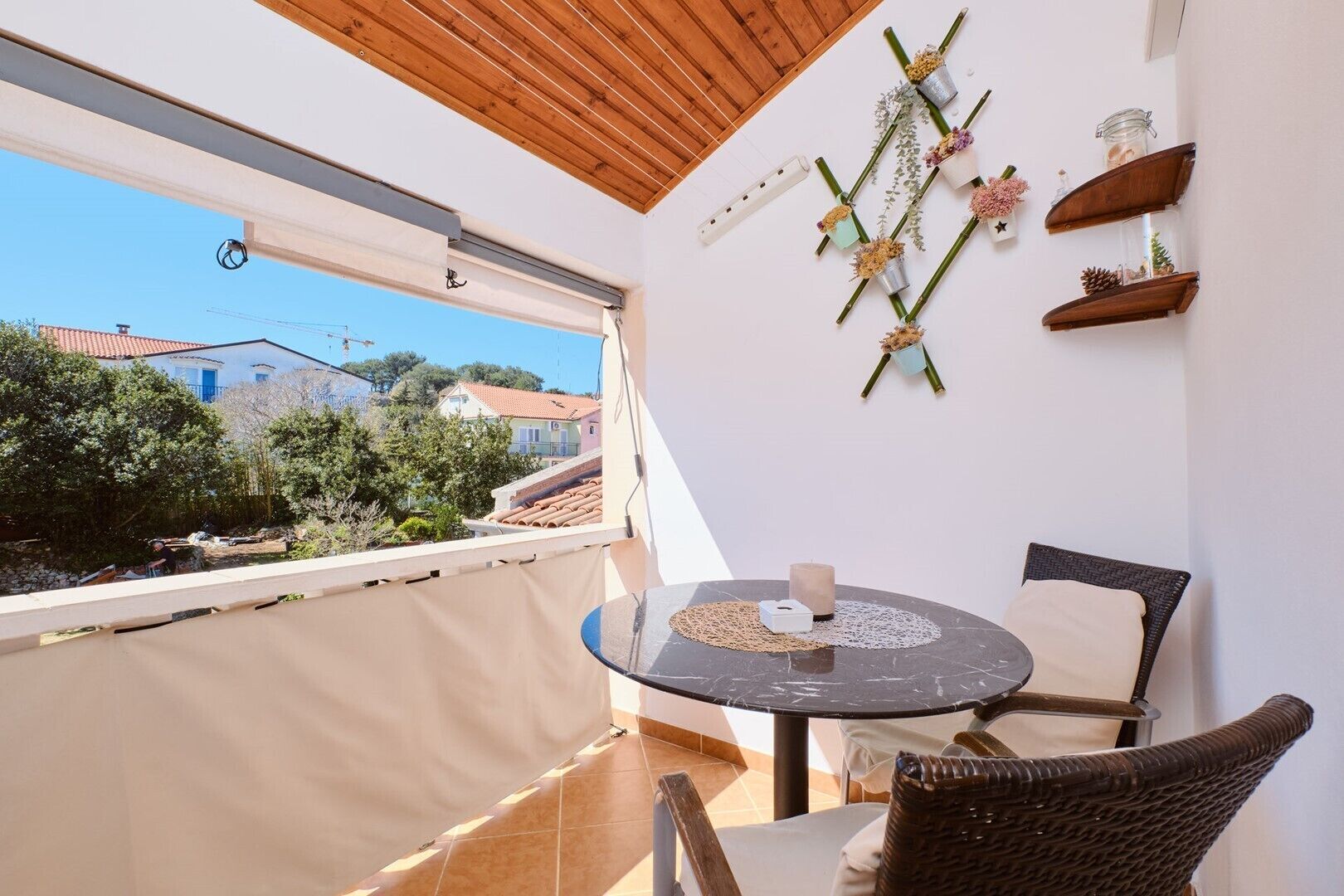 Un balcone con un tavolo rotondo, sedie in vimini, vista su edifici e alberi, e mensole decorative a parete.