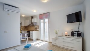 Quarto moderno e bem iluminado, com cozinha compacta, espaço para refeições e TV na parede.