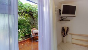 Ein Zimmer mit Holzboden, einem Sessel, einem an der Wand befestigten Fernseher und einem Fenster mit Blick ins Grüne.