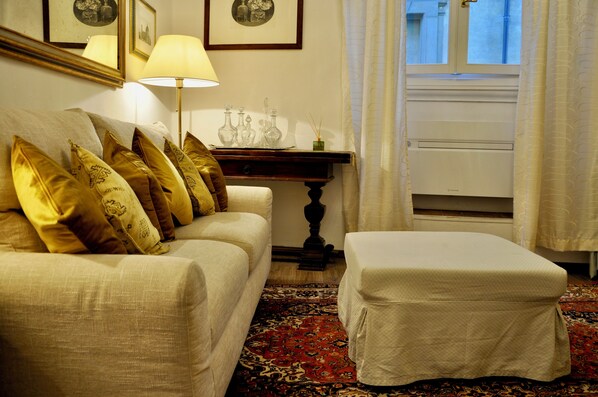 A room with a sofa, an ottoman, a lamp, a small table with decorative items, and a window with curtains.