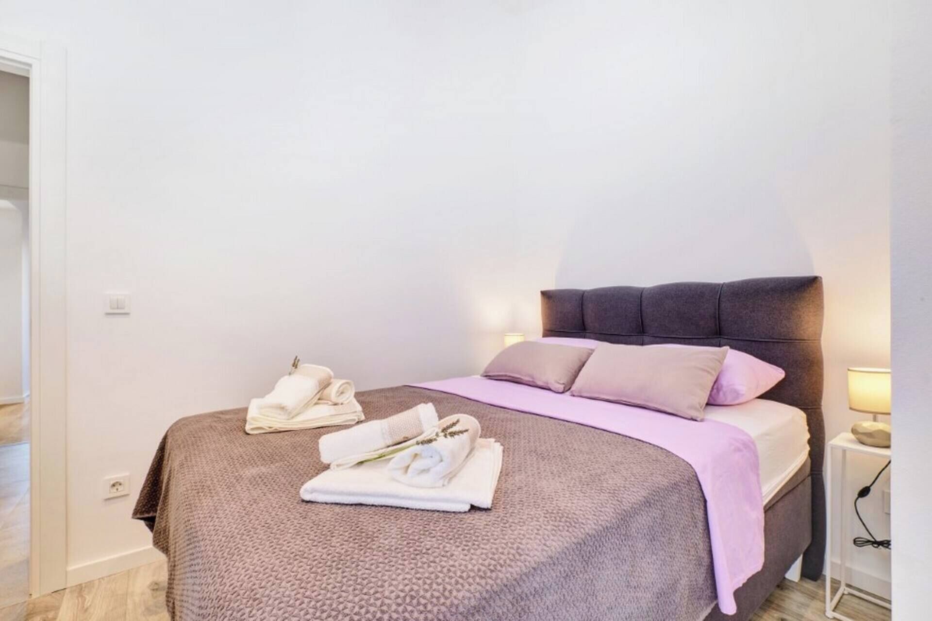 A neatly made bed with a brown headboard, a pink blanket, and two pillows. There are two folded towels on the bed.