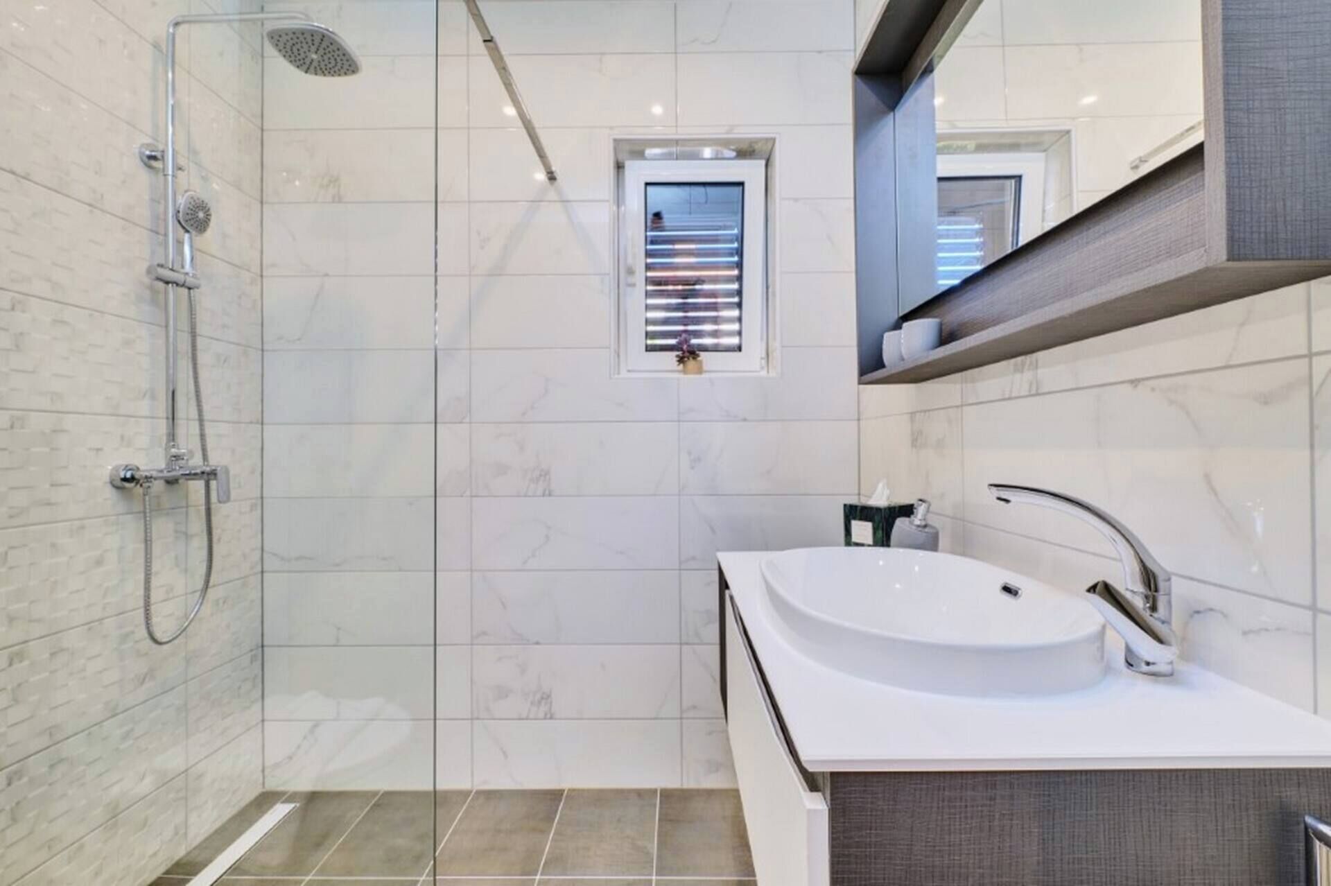 A modern bathroom with a glass-enclosed shower, a white sink, and a small window.
