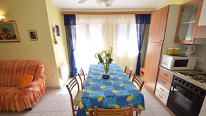 Comedor con una mesa preparada para cuatro, cocina con estufa y microondas, y sofá con un cojín naranja.