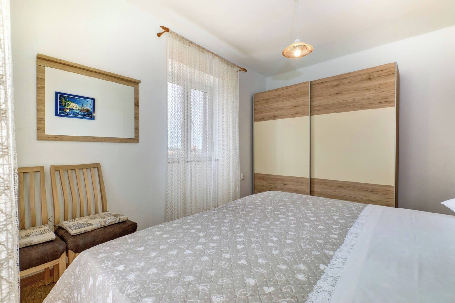 A bedroom with a bed, a wooden wardrobe, a framed picture, and a window with sheer curtains.