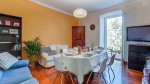 Un comedor con una mesa redonda dispuesta para cuatro personas, rodeada de sillas blancas. Se observa una estantería y una planta en maceta al frente.