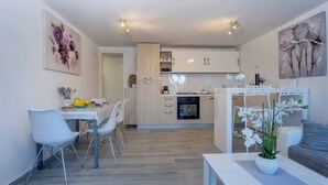 Une cuisine moderne avec des meubles blancs et un four encastré, un coin repas avec une table et des chaises, et un salon avec un canapé et une orchidée en pot.