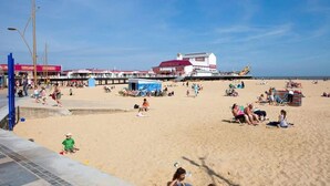 Ein Strand mit badenden und spazierenden Menschen, ein Steg mit Gebäuden und ein klarer Himmel.