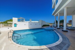 Una piscina en la azotea con cielo azul despejado, un edificio blanco y una palmera al fondo.