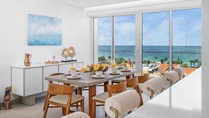 A dining area with a view of the ocean, a white cabinet with decorative items, and a large window.