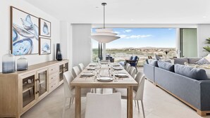 A modern dining area with a wooden table set for six, a grey sofa, and a city view through large windows. - Luxury Coastal Condo with Panoramic Views of Aruba (Noord)