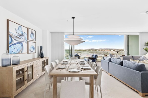 A modern dining area with a wooden table set for six, a grey sofa, and a city view through large windows.