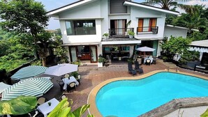 A two-story house with a swimming pool, outdoor seating, and lush greenery.