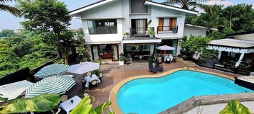 Una casa de dos pisos con piscina, mobiliario de exterior y abundante vegetación.